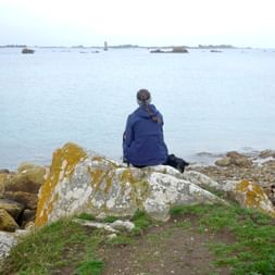 Person in blauer Jacke sitzt auf flechtenbedecktem Felsen mit Blick auf ruhige See mit kleinen Inseln bei Tréguier, Bretagne. Grünes Gras im Vordergrund.