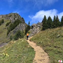 Wanderin in oranger Jacke beim Aufstieg am Stierenberg. Felsige Gipfel und Nadelbäume säumen den Weg unter blauem Himmel.
