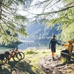 Two cyclists with mountain bikes rest on a wooden bench by a pristine mountain lake surrounded by evergreen trees and mountains.
