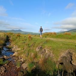 Wanderer geht auf Graspfad durch irische Landschaft mit kleinem Bach, grünen Hügeln und blauem Himmel mit weißen Wolken im Hintergrund.
