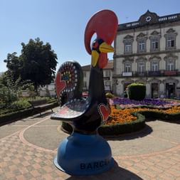 Große verzierte Hahnskulptur auf blauem Sockel mit Aufschrift 'Barcelos' auf Stadtplatz mit bunten Blumenbeeten und historischem Gebäude.