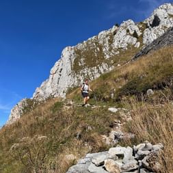 Wanderin mit Rucksack steigt auf felsigem Bergpfad unterhalb weißer Kalksteinfelsen des Stockhorns bei blauem Himmel ab.