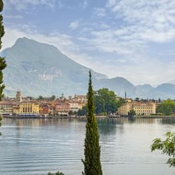 Vue de Riva del Garda au lac de Garde avec des bâtiments colorés le long du front de mer, encadrés de cyprès et de montagnes en arrière-plan.
