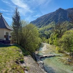 Kleine Kapelle mit Türmchen neben dem klaren Fluss Soča, der durch ein Bergtal bei Mojstrana fließt, umgeben von grünen Bäumen und Bergen.