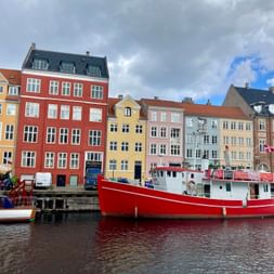 Rotes Fischerboot im Hafen von Kopenhagen vor bunten historischen Gebäuden am Wasser unter bewölktem Himmel.