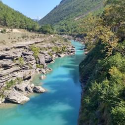 Türkisfarbener Fluss fließt durch felsige Schlucht in Albanien, umgeben von bewaldeten Bergen und Steingebäuden am rechten Ufer.