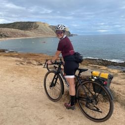 Nina mit Tourenrad auf Küstenfelsen am Strand Praia da Luz. Klippen und ruhiges blaues Meer im Hintergrund unter bewölktem Himmel.