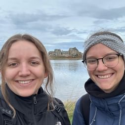 Zwei lächelnde Wanderinnen machen ein Selfie am Le Gouffre de Plougrescant. Hinter ihnen ist ein Haus zwischen Felsen am Wasser sichtbar.
