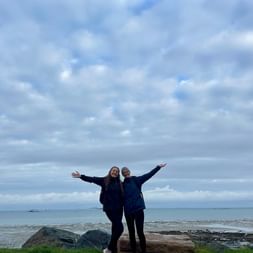 Zwei Frauen mit ausgestreckten Armen stehen auf Gras an der felsigen Küste von Port-l'Épique in der Bretagne unter bewölktem Himmel.