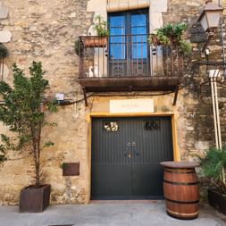 Verwitterte Steinfassade mit blauen Fenstertüren, Eisenbalkon mit Pflanzen, dunkler Holzeingangstür und einem Holzfass neben Topfbäumen.