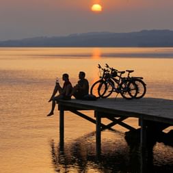 Two bikers sit on the jetty with their bikes, watching a beautiful sunset.