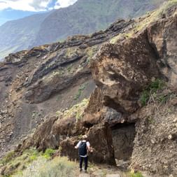 Steiniger Abstieg auf El Hierro Wanderweg Wanderer navigiert steiles felsiges Gelände auf El Hierro mit vulkanischen Felsformationen und Berghängen im Hintergrund unter bewölktem Himmel.