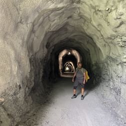 Wanderin mit Rucksack und gelber Tasche in beleuchtetem Felstunnel. Mehrere gewölbte Öffnungen entlang des Weges sichtbar.