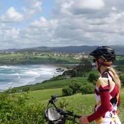 Female cyclist with helmet and colorful jersey overlooking Oyambre beach and coastal landscape with green hills and cloudy sky.