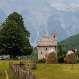 Steinkirche im Balkan-Bergtal Kleine Steinkirche mit Glockenturm und Kreuz in grünem Tal mit Heuballen, großem Baum und dramatischen Berggipfeln im Hintergrund.