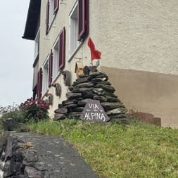 Steinhaufen mit Via Alpina-Schild und Schweizer Flagge neben einem Haus mit roten Fensterläden. Blumen und Gras umgeben die Markierung.