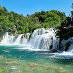 Mehrstufiger Wasserfall, der in türkisfarbene Becken fällt, umgeben von üppigem grünem Wald im Nationalpark Krka, Kroatien.