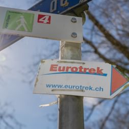 Wegweiser mit Eurotrek-Logo und Wanderwegsmarkierungen vor blauem Himmel mit kahlen Ästen in Courtedoux, Jura, Schweiz.