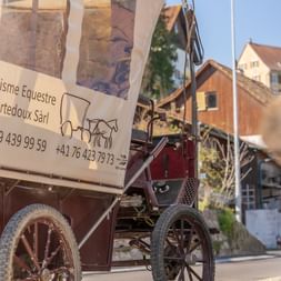 Traditioneller Planwagen mit weißer Plane und Schild 'Tourisme Equestre Courtedoux Sàrl', geparkt in einer Dorfstraße im Jura Ajoie.