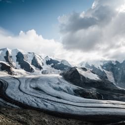 Une image à couper le souffle du glacier de la Diavolezza qui descend en courbe de la montagne enneigée au centre. En haut à droite, il y a des nuages dans différentes nuances de gris qui laissent apparaître un peu de ciel bleu dans le coin supérieur gauche.
