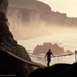 Person überquert die Carrick-a-Rede Hängebrücke in County Antrim mit dramatischen Küstenklippen und nebligem Meeressprühnebel im Hintergrund.