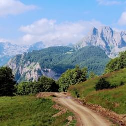 Bergstraße durch Balkan-Landschaft Schotterstraße schlängelt sich durch grüne Wiesen mit dramatischen felsigen Berggipfeln im Hintergrund unter teilweise bewölktem Himmel.