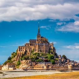 Abbaye du Mont Saint-Michel sur île rocheuse avec bâtiments médiévaux à la base, entourée de plage de sable et de vasières sous ciel bleu nuageux.