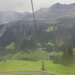 Seilbahngondel über grünen Almwiesen mit bewaldeten Hängen und felsigen Berggipfeln im Hintergrund bei Elm, Schweiz.