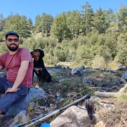 Mann sitzt auf Stein vor Waldlandschaft und ein kleiner Hund lehnt an seinen Rücken