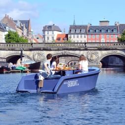 Blaues GoBoat mit Passagieren fährt durch Kopenhagener Kanal vorbei an historischer Steinbrücke und bunten Uferhäusern unter bewölktem Himmel.