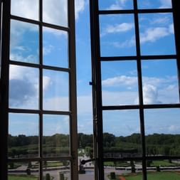 Blick durch hohe Fenster im Schloss Drottningholm auf formale Barockgärten mit geometrischen Rasenflächen, Wegen und Bäumen unter blauem Himmel.
