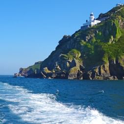 Weißer Leuchtturm auf grünen Klippen der Insel Sark mit felsiger Küste und blauem Meer. Bootsspur im Vordergrund sichtbar.