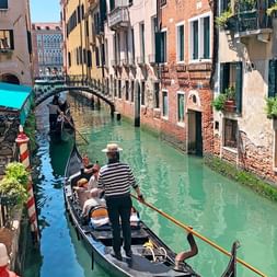 Gondoliere im gestreiften Hemd steuert schwarze Gondel durch schmalen Venedig-Kanal mit historischen Gebäuden, Brücke und Restaurantterrasse.