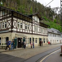 Traditionelles Fachwerkhotel Lichtenhainer Wasserfall mit dunklen Holzbalken und weißen Wänden am Fuße eines bewaldeten Hanges.