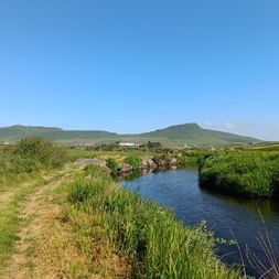 Path beside a river in Dingle, Ireland, in bright sunshine. Green hills in the background.