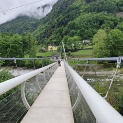 Man sieht wie eine Velofahrerin die Hängebrücke bei Maggia überquert. Im Hintergrund ist in Mitten von Bäumen ein kleines Dorf erkennbar.