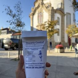 Hand hält weiße Papiertüte der Bäckerei Doçaria Central in Arcos de Valdevez, mit historischer Kirche und blauem Himmel im Hintergrund.
