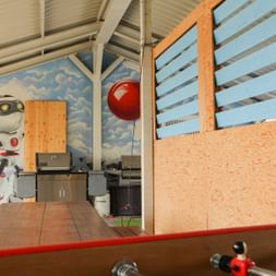 Moderner Büro-Pausenraum mit sichtbaren Deckenbalken, himmelbemalter Wand, Dartscheibe, Tischfußball, Küchenbereich und rotem Ballon.