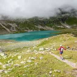 Trail runner running in the direction of a mountain lake. Trailrunning Via Grischuna. Hiking vacation with Eurotrek.