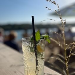 Glas mit Eisgetränk, Minzblättern und schwarzem Strohhalm auf Holztisch. Unscharfer Küstenhintergrund mit Hafen und blauem Himmel in Cornwall.