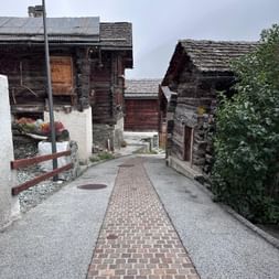 Schmaler Kopfsteinpflasterweg zwischen traditionellen dunklen Walliser Holzchalets in Ayer. Alte verwitterte Gebäude mit Steindächern unter bewölktem Himmel.