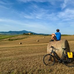 Homme en chemise bleue assis sur une grande botte de foin dans la campagne toscane. Vélo noir avec sacoches jaunes à côté.