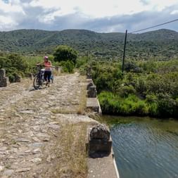 Radfahrerin mit Kinderanhänger auf Steinbrücke Radfahrerin mit Kinderanhänger überquert alte Steinbrücke ohne Geländer über grünem Wasser, umgeben von Hügeln und Vegetation.