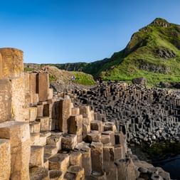 Sechseckige Basaltsäulen am Giant's Causeway mit grünen Küstenhügeln und blauem Himmel. Besucher zwischen den dunklen Steinformationen am Wasser.