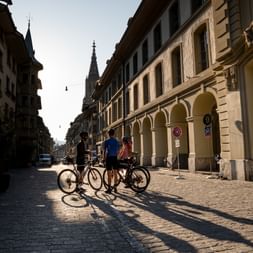 Three cyclists with bikes standing on a cobblestone street in Bern's old town, with historic buildings and a church spire in the background.
