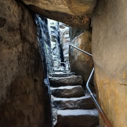 Enge Steintreppe mit Metallgeländer führt aufwärts durch einen engen Felsdurchgang am Pfaffenstein, mit Licht am oberen Ende sichtbar.