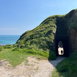 Steintunnel durch einen grasbewachsenen Küstenhügel in Cornwall. Eine Person geht durch den Tunnel zum Strand. Blaues Meer links sichtbar.