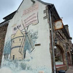 Weißes Gebäude in Perros-Guirec mit gemaltem Windmühlen-Wandbild. Schild 'Ti Loz Patisserie Boulangerie Chocolatier' über dem Kunstwerk.