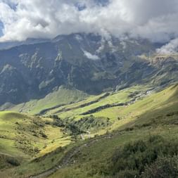 Grünes Alpental mit sanften Hügeln und Bach, umgeben von dramatischen Berggipfeln, die teilweise von Wolken bedeckt sind, in der Ostschweiz.
