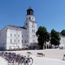 Weißes barockes Salzburg Museum mit markanten Uhrenturm auf sonnigem Platz. Fahrräder stehen davor, Bänke säumen den Platz.